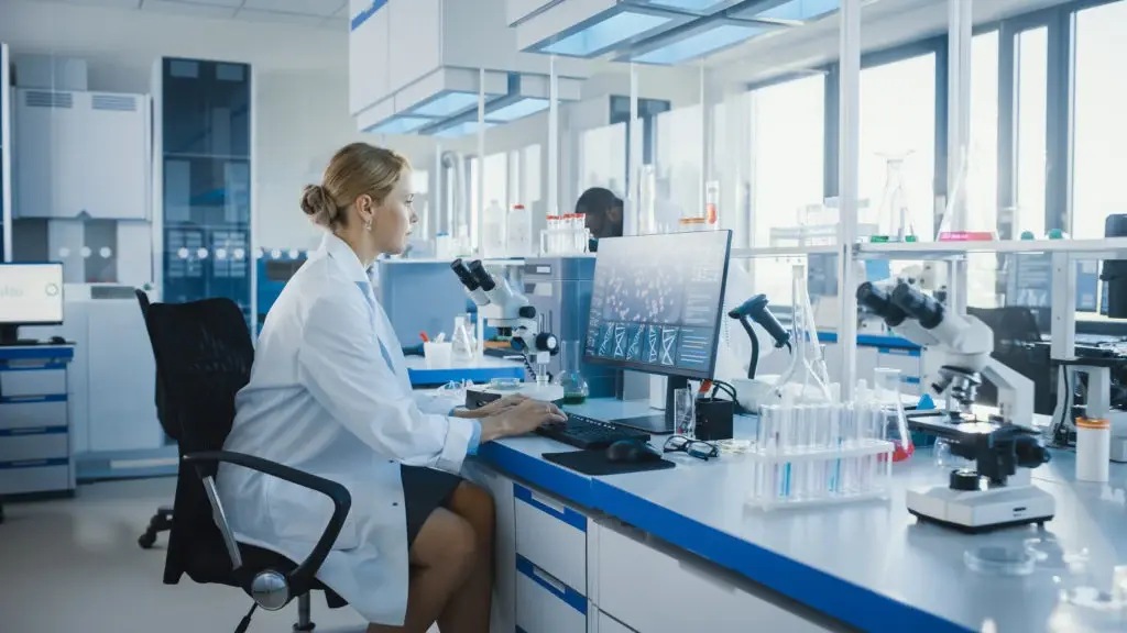 Female Microbiologist Working on Computer with Display Showing Gene Editing Interface and Using Microscope
